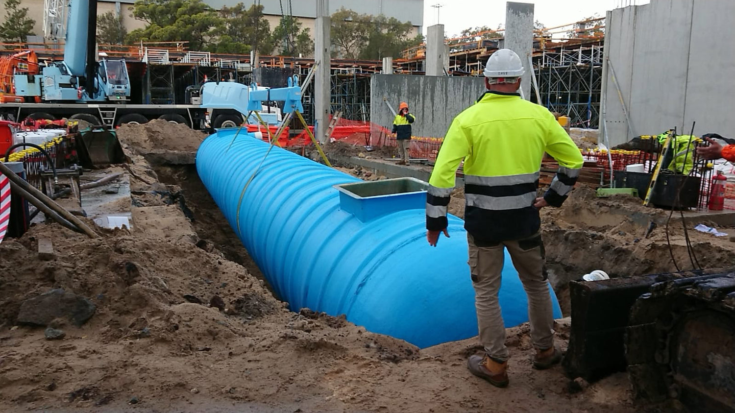A construction worker in high-visibility gear overseeing a large blue underground liquid storage tank situated in an excavation trench.