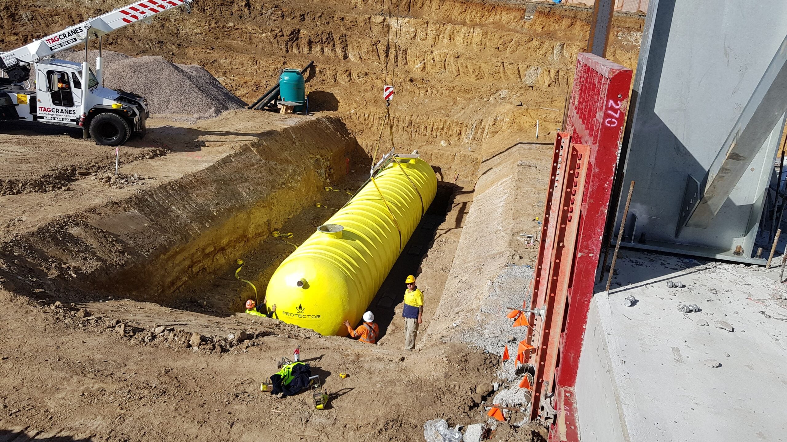 Construction workers guiding a large yellow Hydroprotector underground tank being lowered into a dirt trench by a crane.