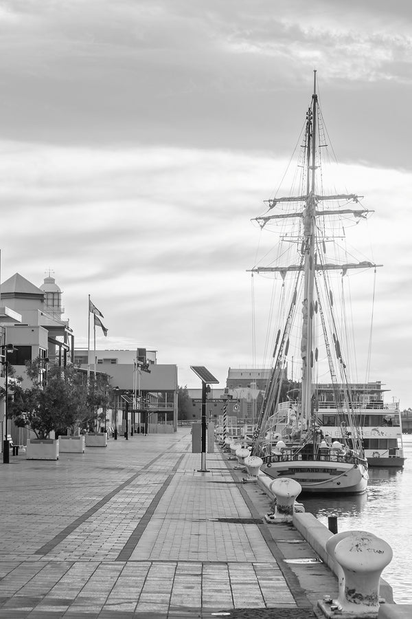 Black and white photograph of a tall sailing ship moored beside a waterfront boardwalk.