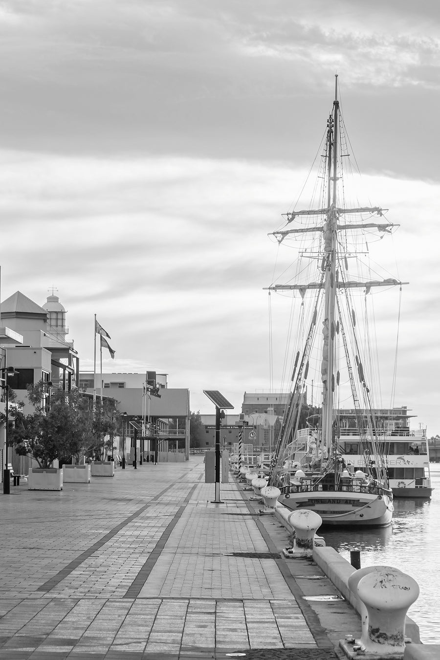Black and white photograph of a tall sailing ship moored beside a waterfront boardwalk.