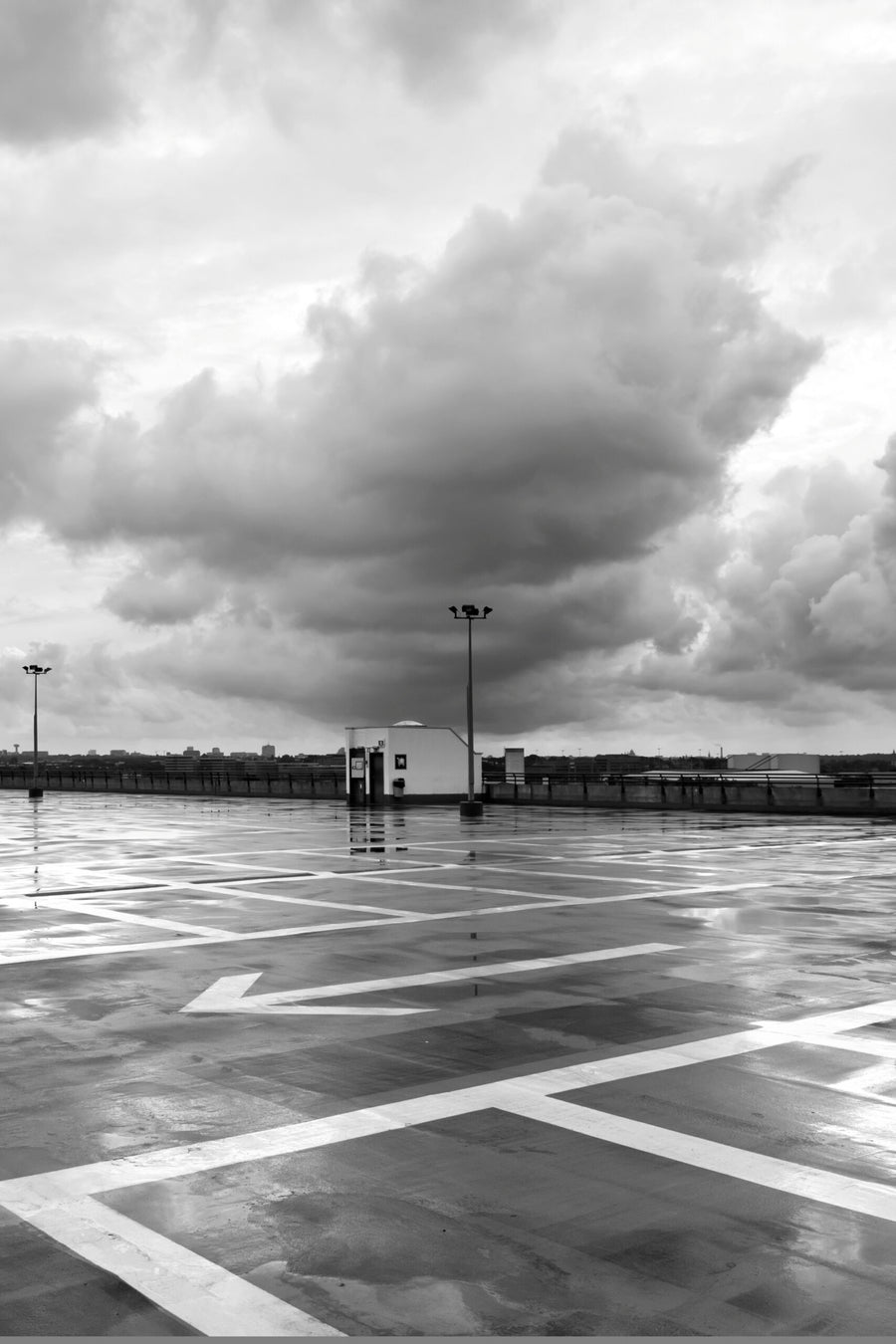 Black and white photograph of an empty, wet rooftop parking lot under a dramatic, cloudy storm sky.