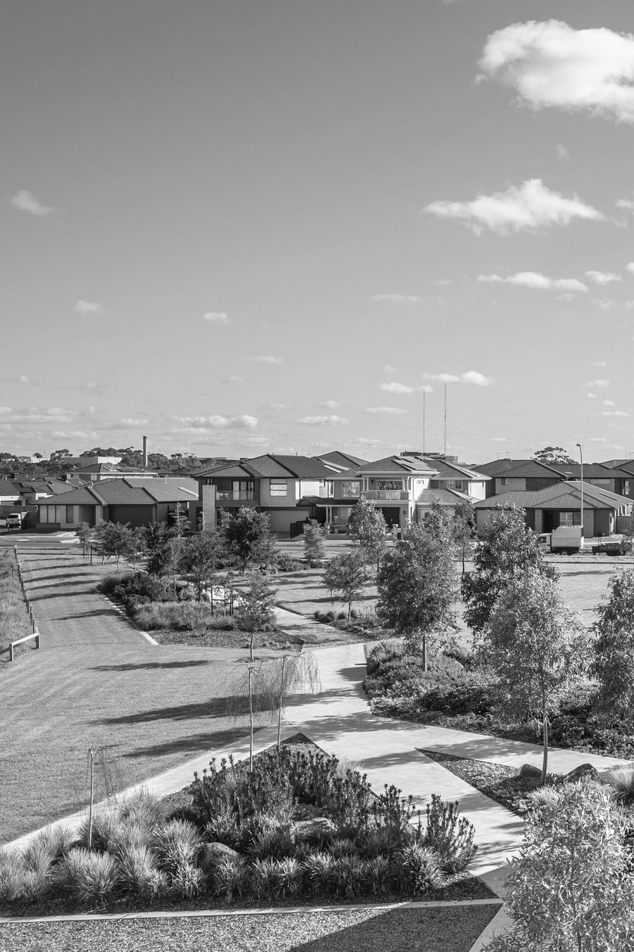Black and white photograph of a suburban neighborhood park with walking paths, trees, and houses under a partly cloudy sky.